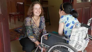 Die Freiwillige sitzt hockend vor einem Kind in einem Rollstuhl und lacht das Kind an.